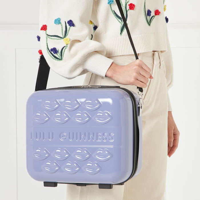A purple Lulu Guinness makeup case with a black strap, decorated with embossed lips, being held by a person wearing a cream-colored sweater.