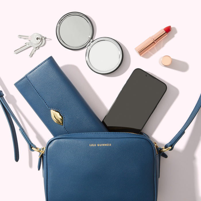 A navy blue Lulu Guinness crossbody bag with gold hardware, containing a smartphone, wallet, compact mirror, lipstick, and keys, all arranged on a white background.