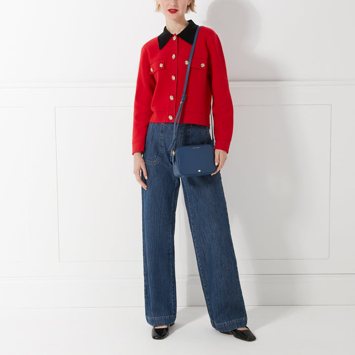 A woman in a red cardigan and blue jeans is carrying a navy blue crossbody bag against a white background.