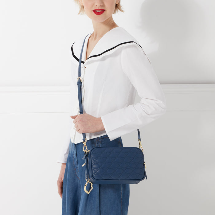 A woman in a white collared shirt and jeans carries a navy blue quilted crossbody bag with a gold clasp. The background is white.