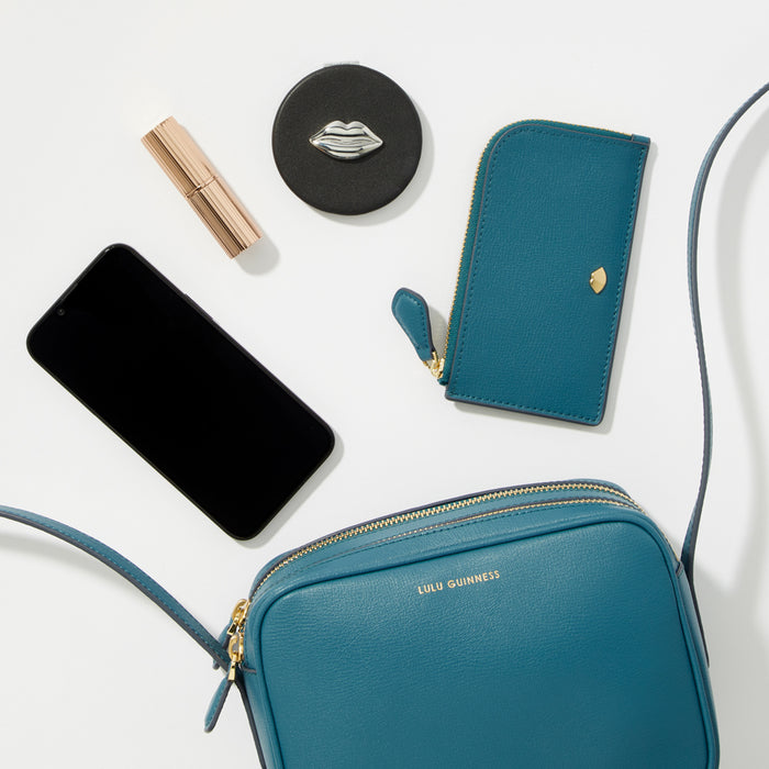 Teal crossbody bag, smartphone, lipstick, compact mirror with lip design on white background.