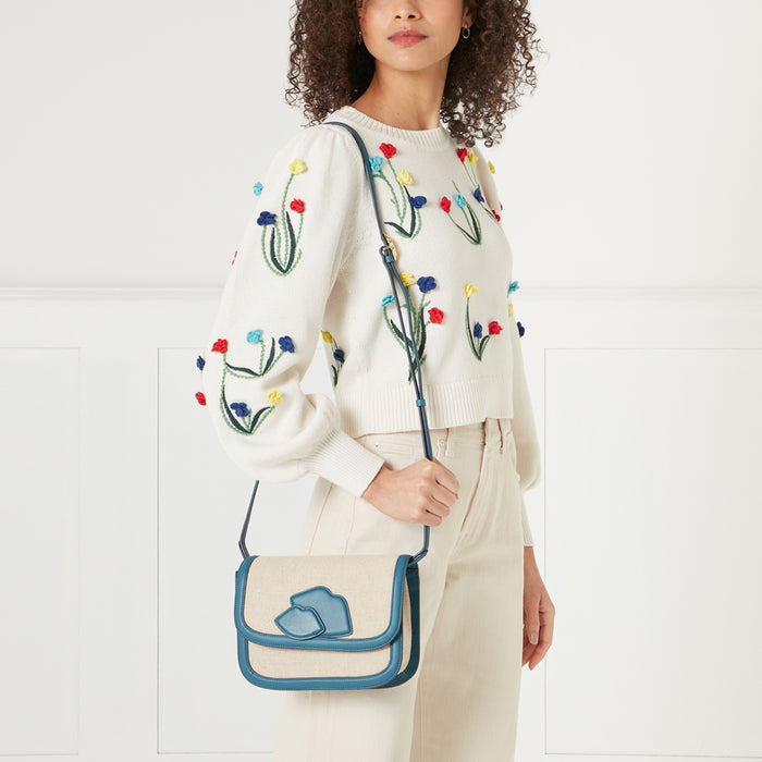 Woman in cream floral sweater holding a teal and beige crossbody bag with lip detail against a white background.