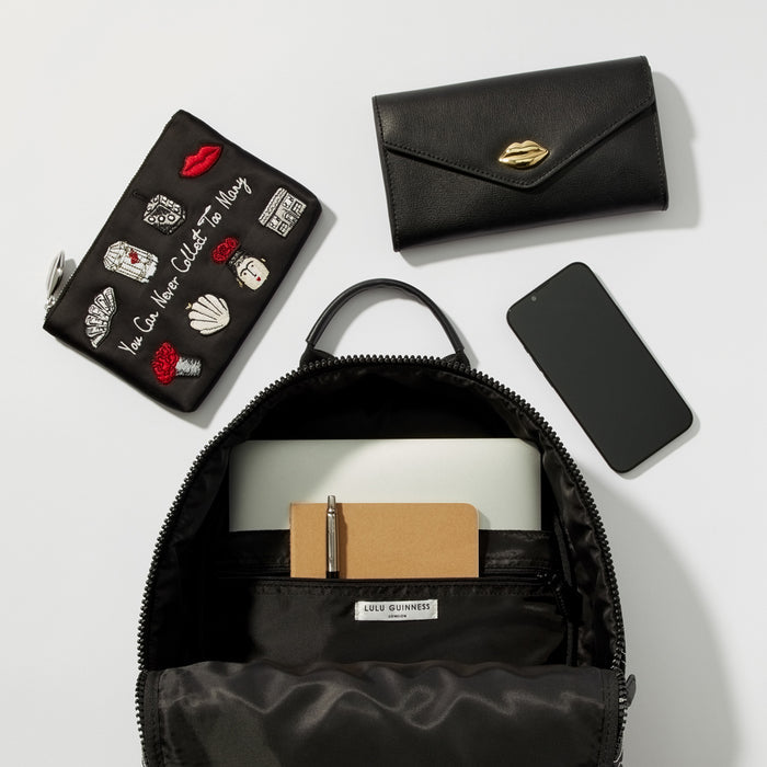A black Lulu Guinness backpack is open to reveal its interior, which contains a laptop, notebook, and pen. A black leather wallet and a small black makeup bag with embroidered details sit beside the backpack along with a black smartphone, all on a white background.
