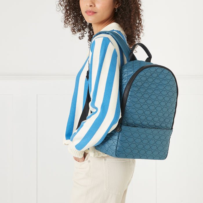 Woman wearing a blue and white striped sweater and beige pants, modelling a teal quilted backpack with a lip print pattern.