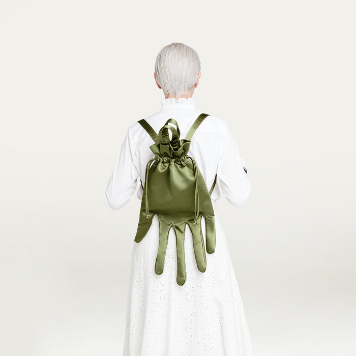 Person wearing a white outfit with a green satin hand-shaped backpack on a light gray background