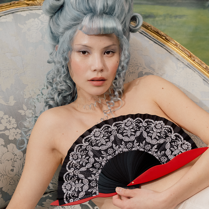 Woman with styled hair holding a black lace fan in an ornate room.