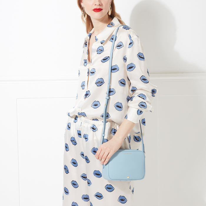 Woman in a white silk pyjama set with a blue lip print holds a pale blue crossbody bag against a white background.