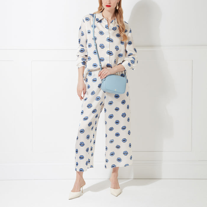 A woman in a white lip-print jumpsuit carries a light blue crossbody bag against a white background.