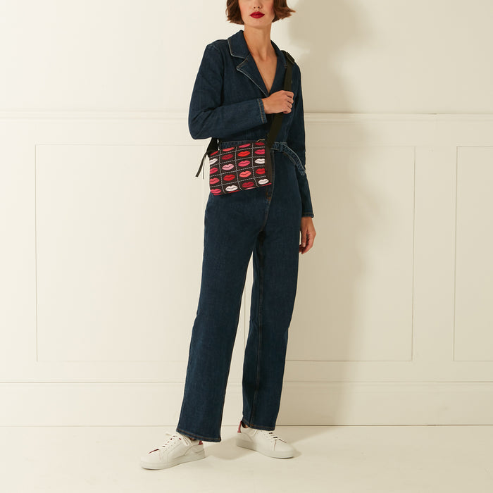 Woman in dark wash denim jumpsuit wearing white sneakers and a crossbody bag with a lipstick print against a white background.