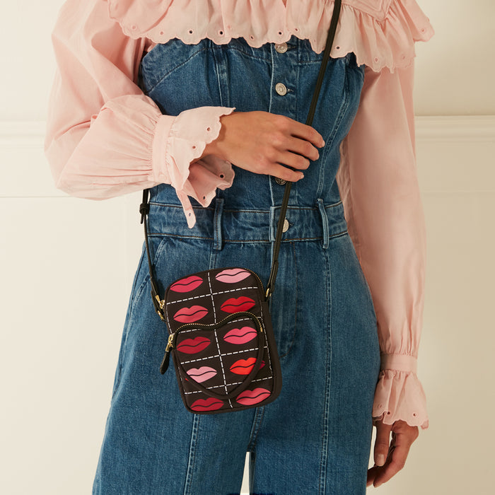 A dark brown crossbody bag with a pink and red lips pattern worn over a denim jumpsuit and a pink eyelet blouse.
