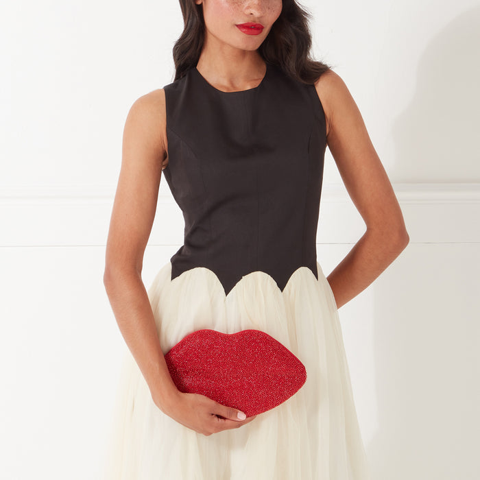 Woman in black and white dress holding a red beaded lip-shaped clutch against a white background.