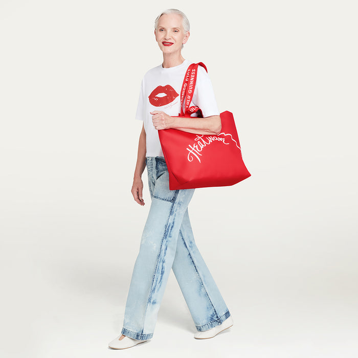 A woman modeling a large red tote bag with white script lettering reading 'Heatwave', styled with a white graphic t-shirt and light-wash jeans against a plain light background.