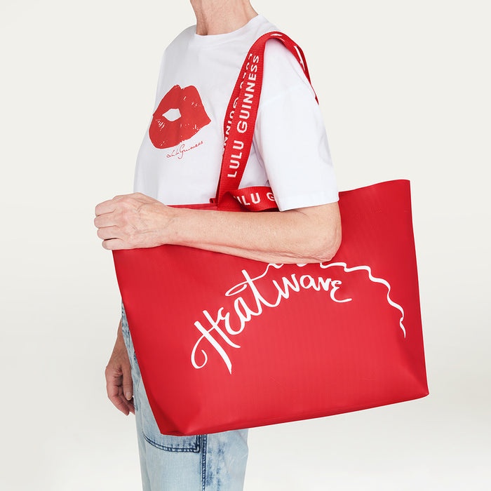 A person wearing a white graphic T-shirt and light blue jeans carries a large red tote bag with a white script logo 'Heatwave' and a red shoulder strap branded with 'LULU GUINNESS'.