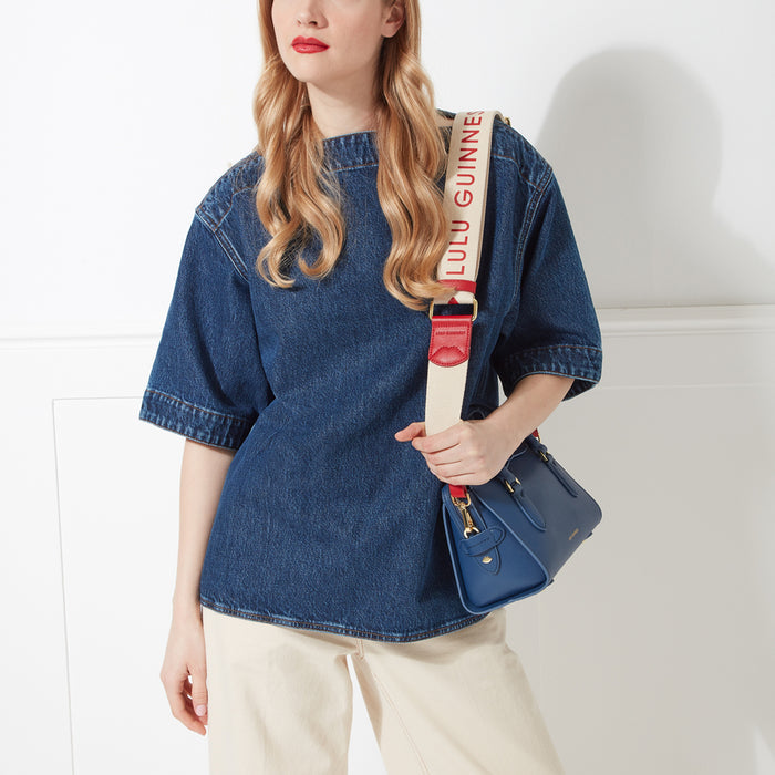 Woman with blonde hair wearing a denim shirt and carrying a navy blue Lulu Guinness crossbody bag with a cream and red strap.