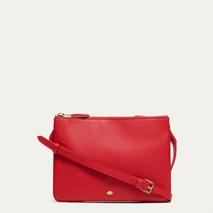 Red crossbody bag with a gold buckle and lip shaped pin on the front on a light gray background