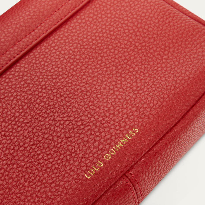 A close-up of a red pebbled leather item with gold foil 'LULU GUINNESS' branding, showing the textured surface and corner detail against a white background.