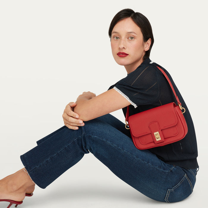 A women in a navy knit top and blue denim jeans sitting on the floor with a red leather bag on her shoulder.