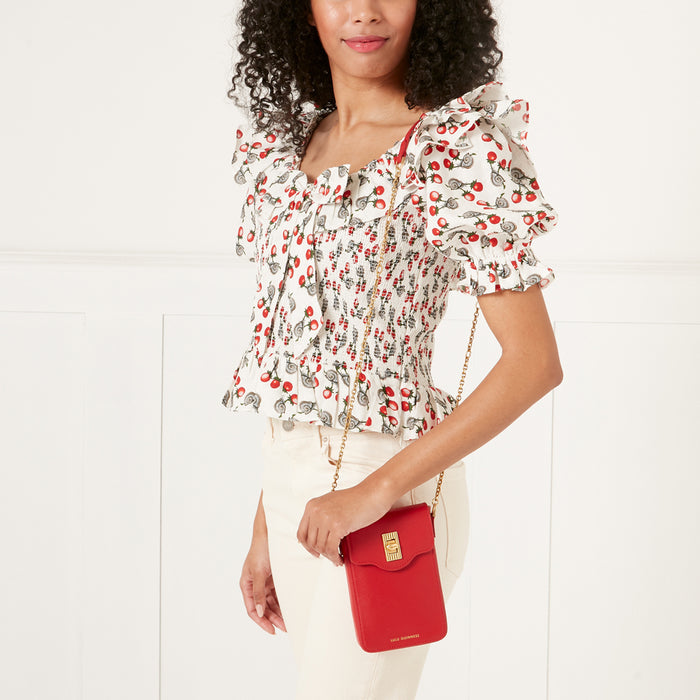 Woman in a cherry-print top holding a red Lulu Guinness phone bag with gold chain strap against a white background.