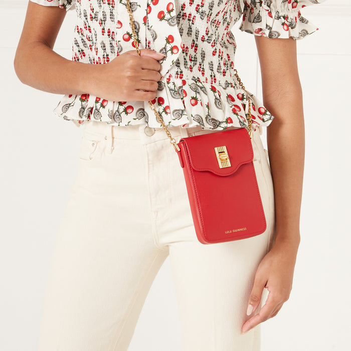 A woman is wearing a red Lulu Guinness crossbody bag on her left shoulder. The bag is small and rectangular. She is wearing cream colored pants and a white top with a red and white snail print.