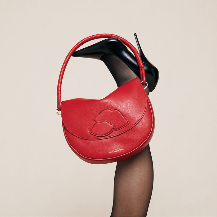 Red handbag with a unique lip design held by a black high-heeled shoe on a beige background