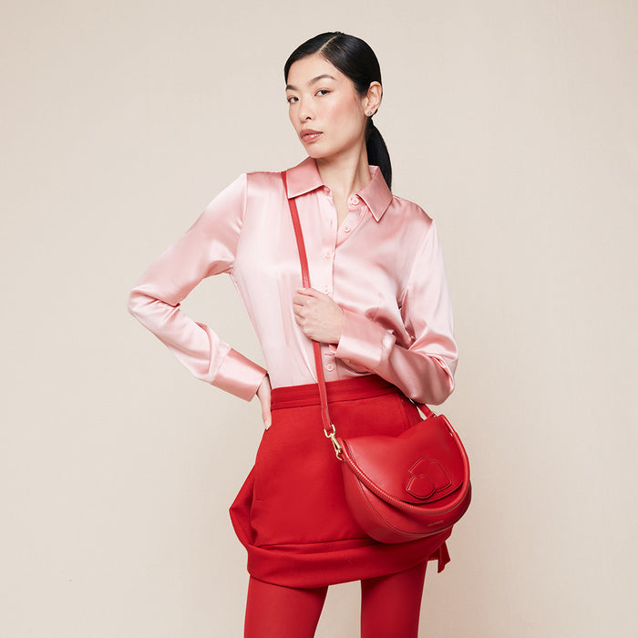 Woman wearing a pink satin shirt and red skirt holding a red leather handbag with lip design on the front against a beige background