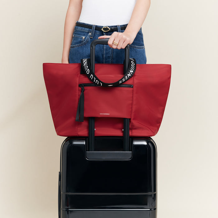 Red tote bag with 'Lulu Guinness' branding held over a black suitcase on a beige background