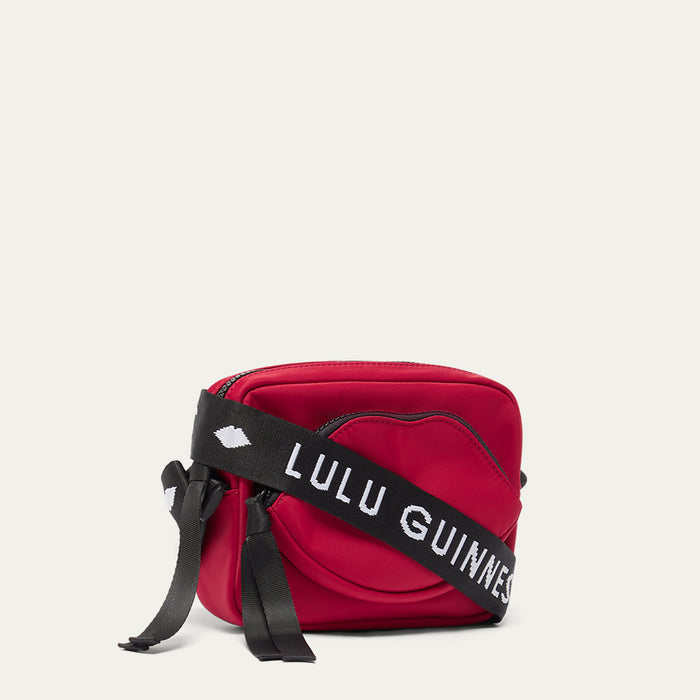 Red crossbody bag with a black strap featuring 'Lulu Guinness' and a lip-shaped pocket on a light grey background