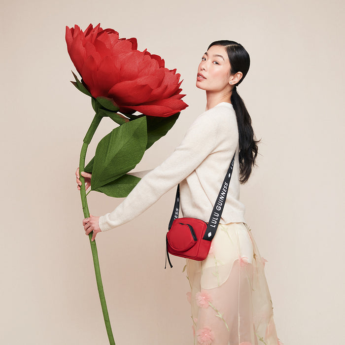 Women holding a large red flower and wearing a red nylon crossbody bag with a 'Lulu Guinness' branded strap