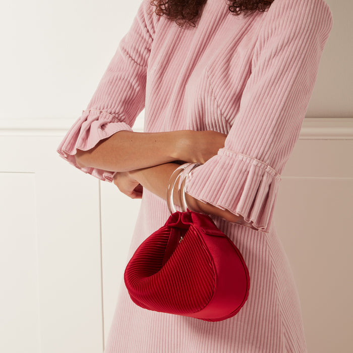A woman in a pink corduroy dress holds a red pleated satin handbag with a clear acrylic handle.