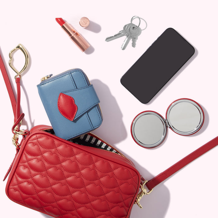 A red quilted crossbody bag with a blue wallet, lipstick, keys, phone, and compact mirror on a white background.