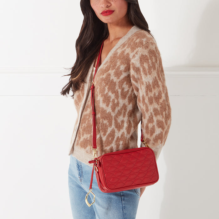 A woman in a beige cardigan and jeans is modelling a red quilted crossbody bag with a gold lip charm. The background is white.