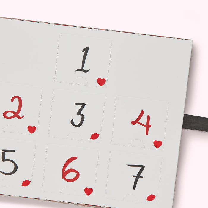 Close-up of a light grey advent calendar with numbers 1-7, each with a small red heart or lip beside it. The background of the calendar has a subtle patterned design.