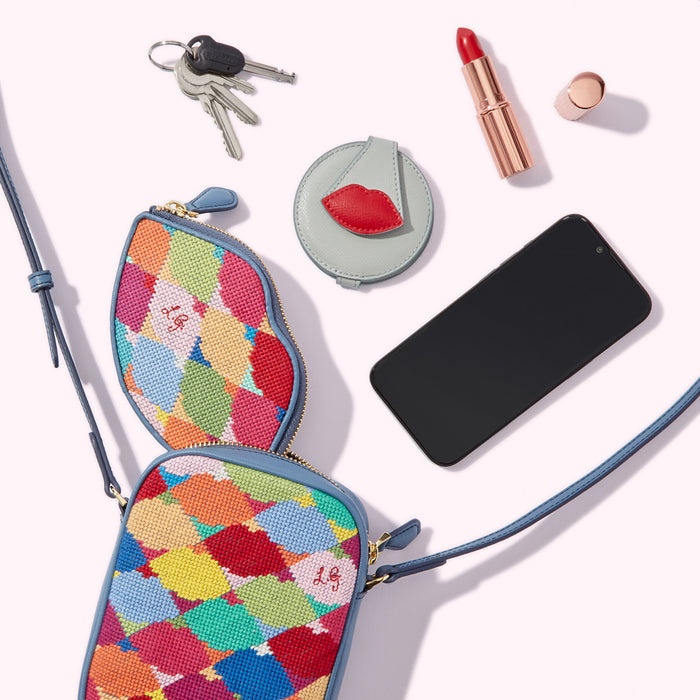 A colourful needlepoint crossbody bag and matching coin purse, a compact mirror, keys, lipstick, and a smartphone arranged on a white background.