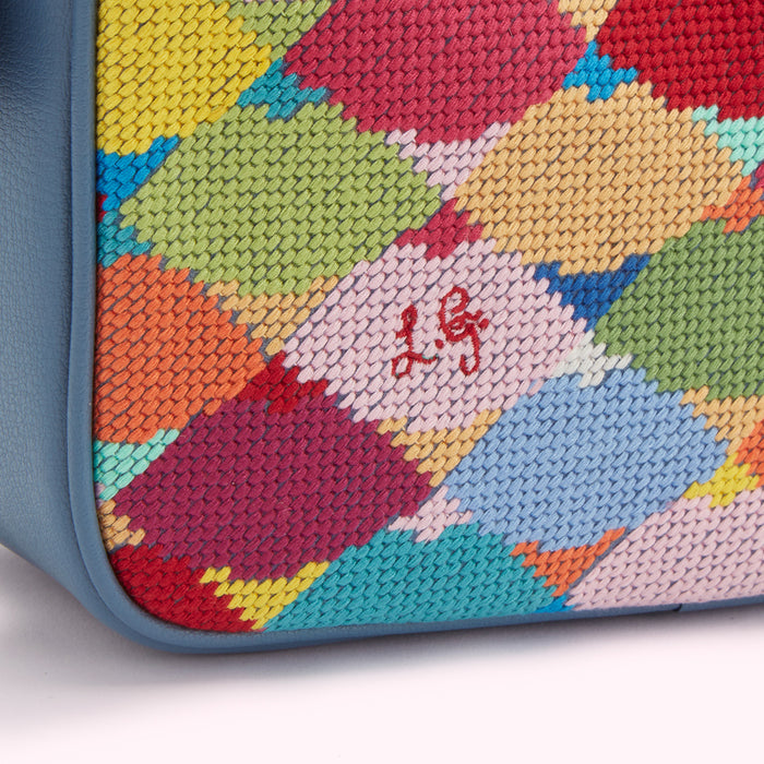 Close-up of a multi-coloured needlepoint textile with the initials "L.G." embroidered in red. The textile is bordered with dark blue leather.