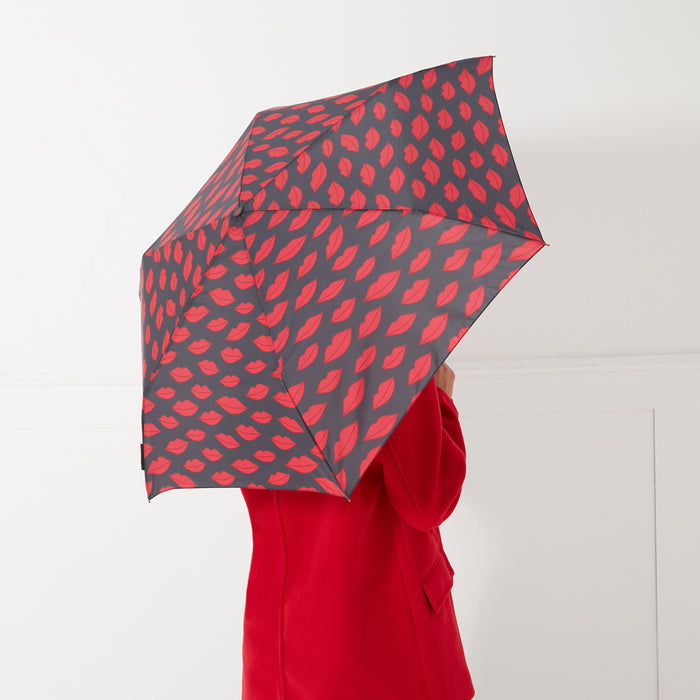 A woman in a red coat holds a dark grey umbrella with a pattern of red lips printed on it.