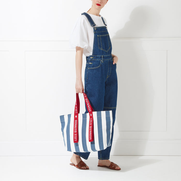 A woman in denim overalls holds a large blue and white striped tote bag with red Lulu Guinness logo straps against a white background.