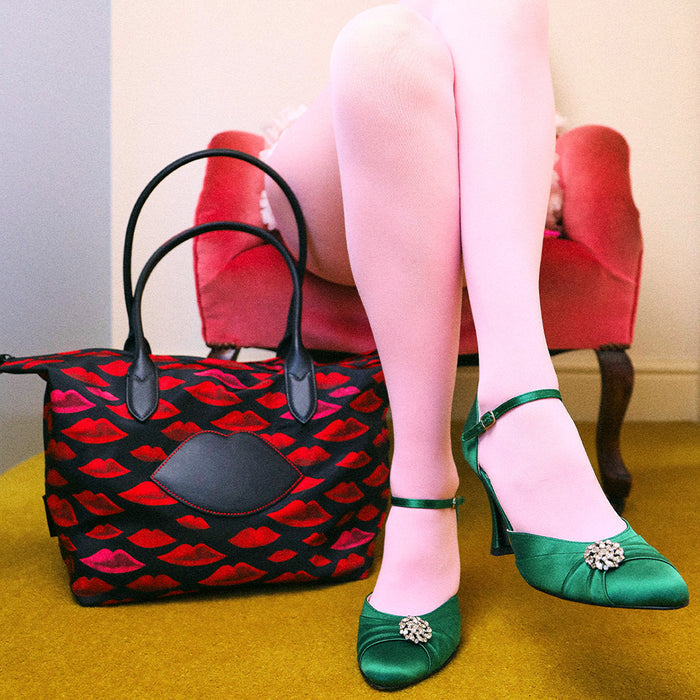 Person wearing green shoes with a brooch, sitting on a red chair next to a red and black patterned handbag.