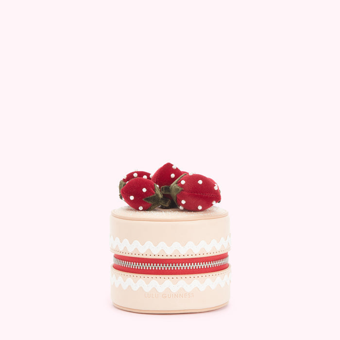 Cream-colored cylindrical purse shaped like a cake, decorated with red velvet strawberries and a red zipper, against a light pink background. The brand "Lulu Guinness" is visible on the purse.