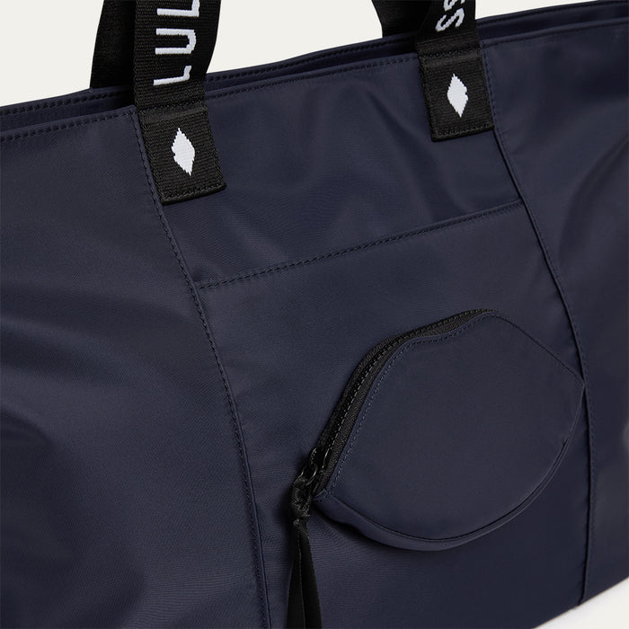 Navy blue tote bag with a lip-shaped pocket and black straps on a light grey background