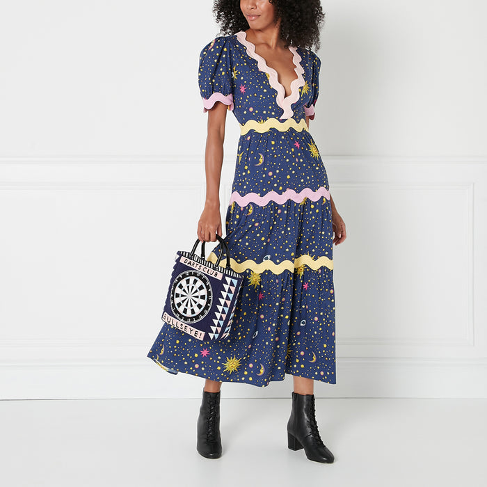 Woman in a navy blue midi dress with a celestial print and pink and yellow details carries a navy tote bag with a dartboard design. The background is white.