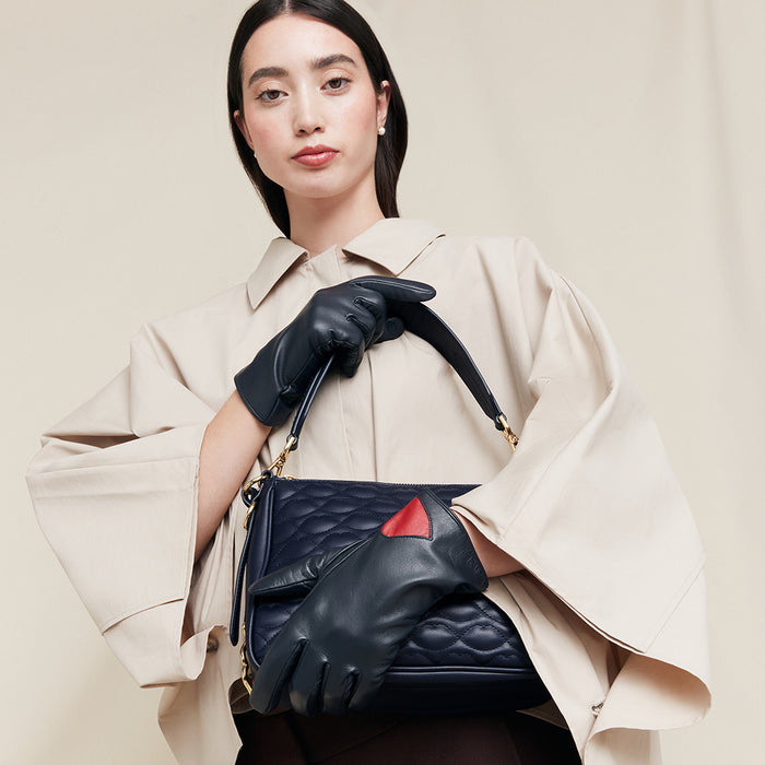 Woman holding a navy leather handbag wearing navy leather gloves with red accents, wearing a beige coat on a beige background