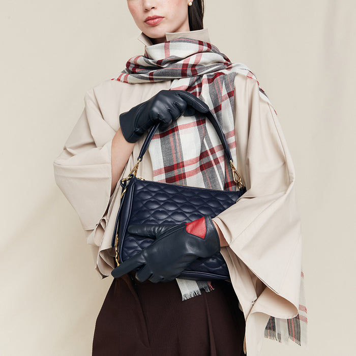 Person holding a navy quilted handbag wearing navy leather gloves with a red accent, wearing a beige coat and plaid scarf.