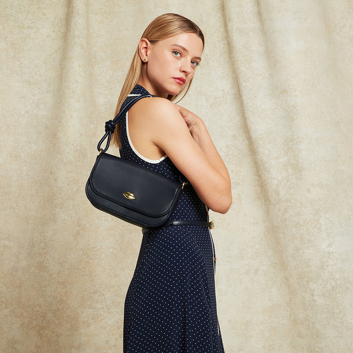 Woman wearing a navy polka dot dress and holding a navy leather shoulder bag against a beige curtain background