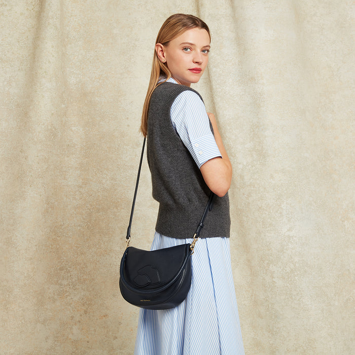 Woman carrying a navy leather handbag showing the crossbody strap against a beige curtain background