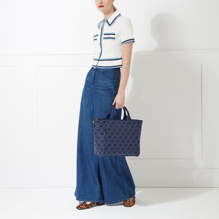Woman in white cropped cardigan and wide-leg jeans holding a navy blue tote bag with a lip print against a white background.
