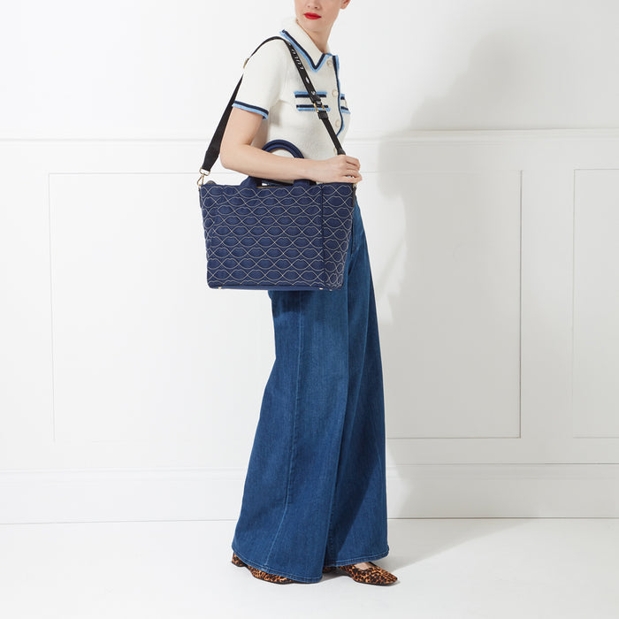 Woman in a white sweater and blue jeans carrying a navy blue and white tote bag against a white wall.