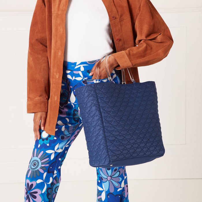 A navy blue quilted tote bag with clear handles is held by a person wearing a brown suede jacket and blue floral pants against a white background.