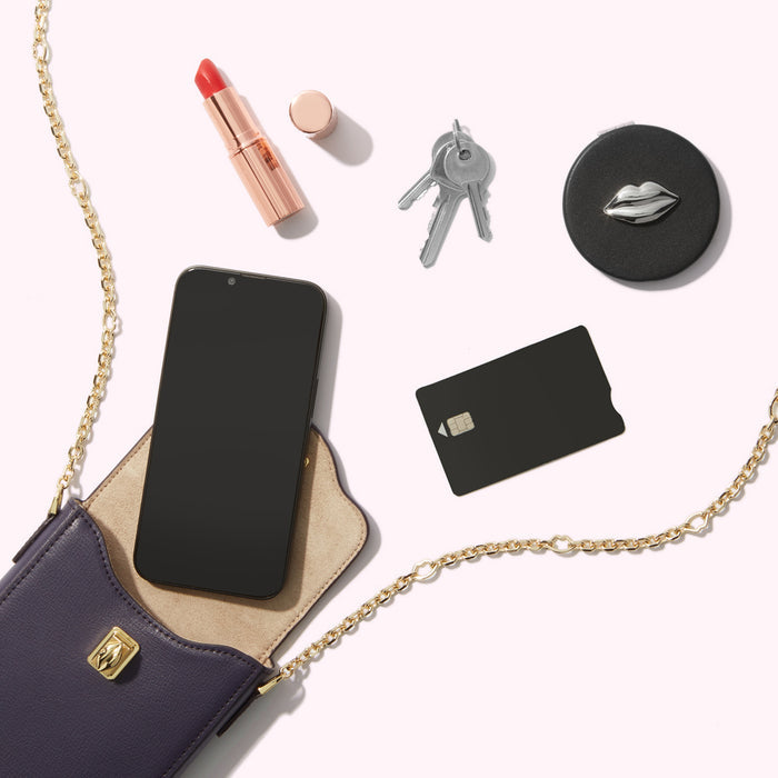 A flatlay of a purple crossbody bag, a smartphone, lipstick, keys, a credit card, and a compact mirror on a white background.