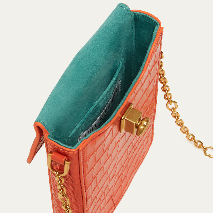 Close-up of a small handbag in textured orange leather with a bright teal interior lining and gold-tone chain strap on a light gray background.