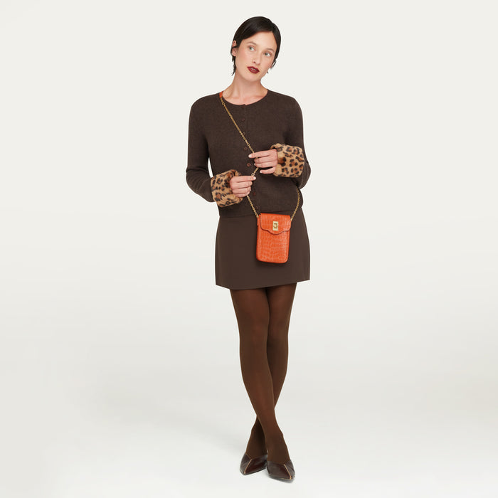 A woman modeling a brown knit mini dress with leopard-print elbow cuffs, wearing brown tights and dark pointed shoes, while carrying a small orange croc-effect embossed leather crossbody bag with a gold chain strap against a light gray studio background.